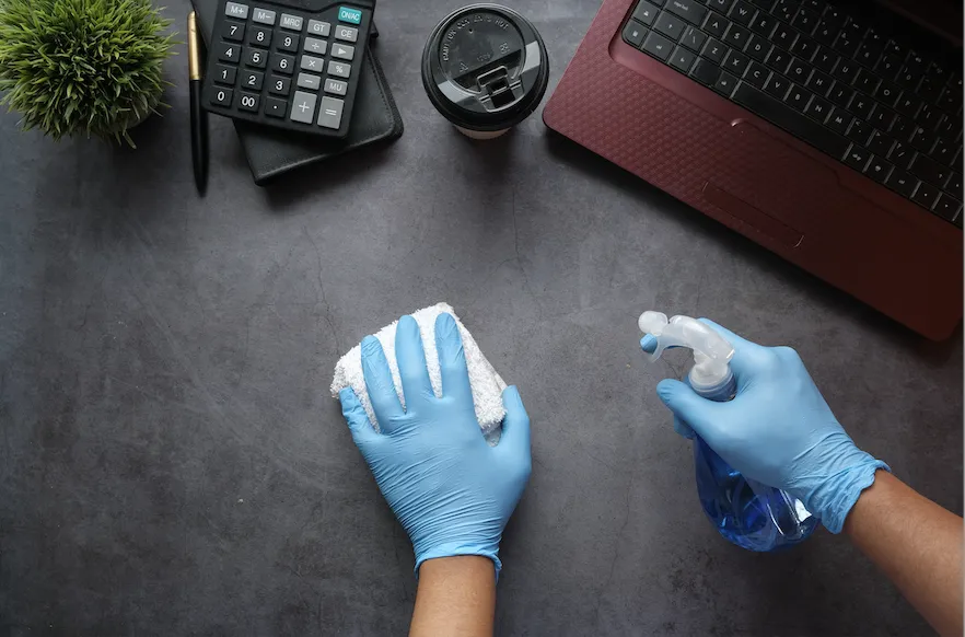 Hands wearing blue latex gloves and using cleaning supplies to clean a desk